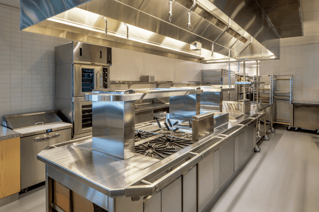 Commercial kitchen with a large stainless steel cooking range, integrated grill section, and overhead ventilation hood; surrounded by ovens, prep counters, and storage racks in a clean, tiled environment designed for high-volume culinary efficiency