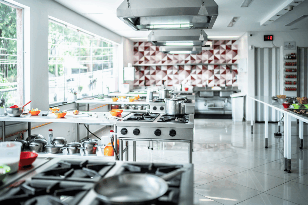 Contemporary commercial kitchen with multiple stainless steel cooking stations, natural light streaming through large windows, and a bold red-and-white geometric tile wall; an ergonomic space blending visual flair with functional design for collaborative culinary work.