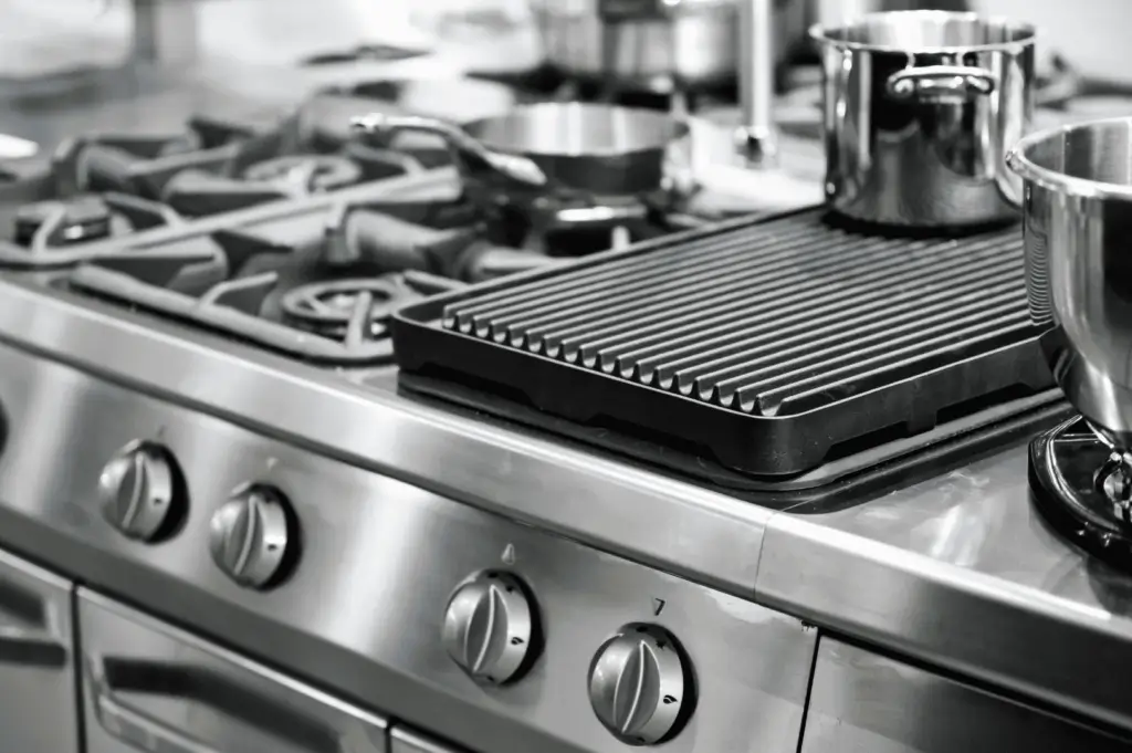 Close-up of a professional-grade stainless steel stove with multiple burners, control knobs, and a cast iron grill pan spanning two burners; a tactile, high-performance setup emphasizing precision and accessibility in commercial cooking environments.