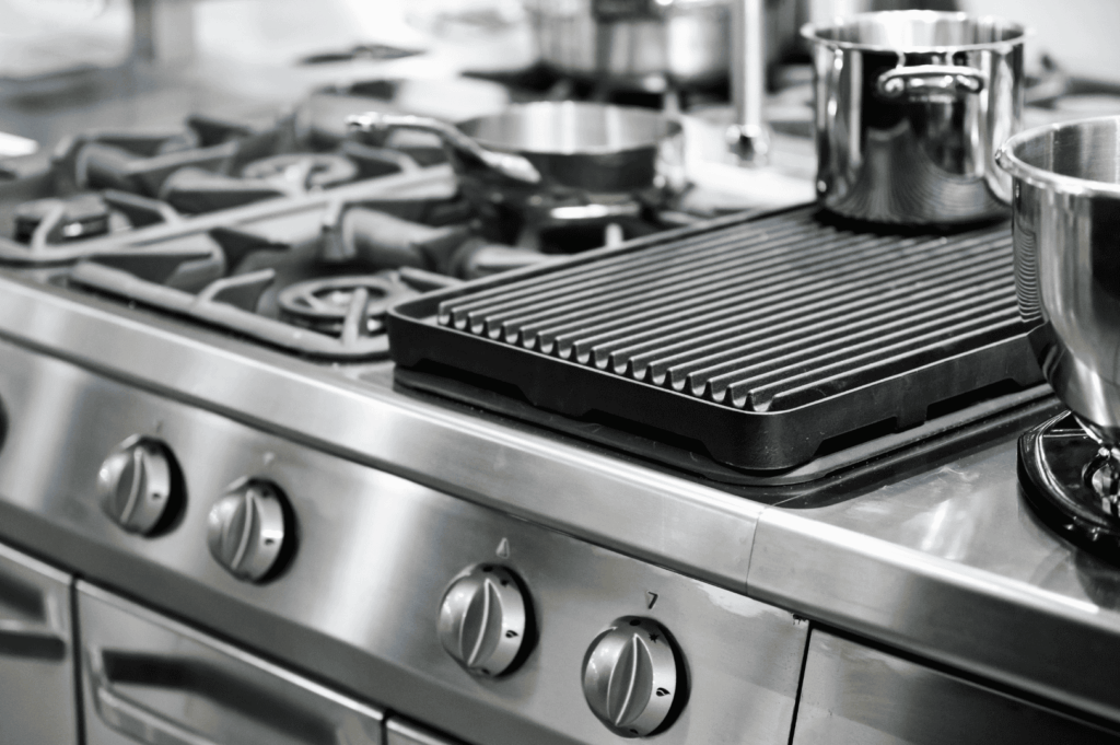 Close-up of a professional-grade stainless steel stove with multiple burners, control knobs, and a cast iron grill pan spanning two burners; a tactile, high-performance setup emphasizing precision and accessibility in commercial cooking environments.