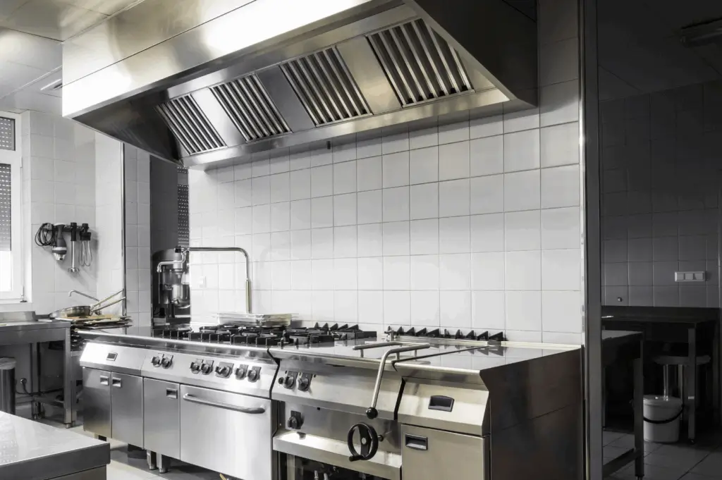 Industrial-grade commercial kitchen with a large stainless steel cooking range, overhead ventilation hood, and tiled walls; a clean, well-lit space engineered for high-volume food prep and ergonomic efficiency