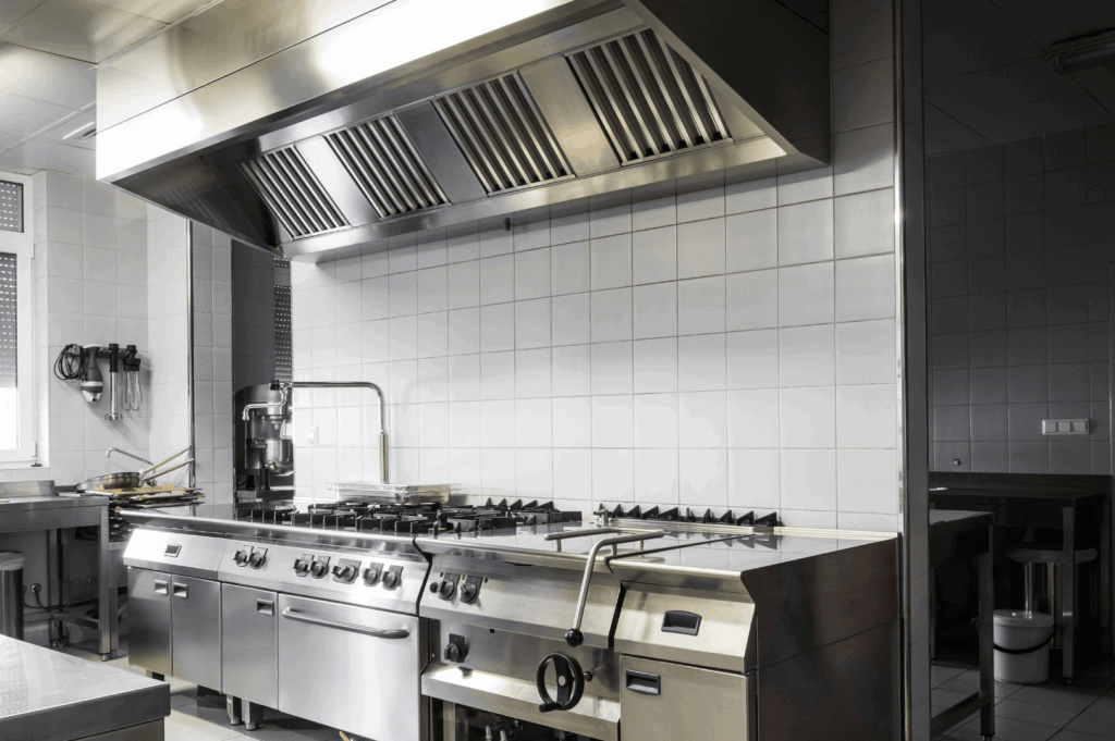 Industrial-grade commercial kitchen with a large stainless steel cooking range, overhead ventilation hood, and tiled walls; a clean, well-lit space engineered for high-volume food prep and ergonomic efficiency
