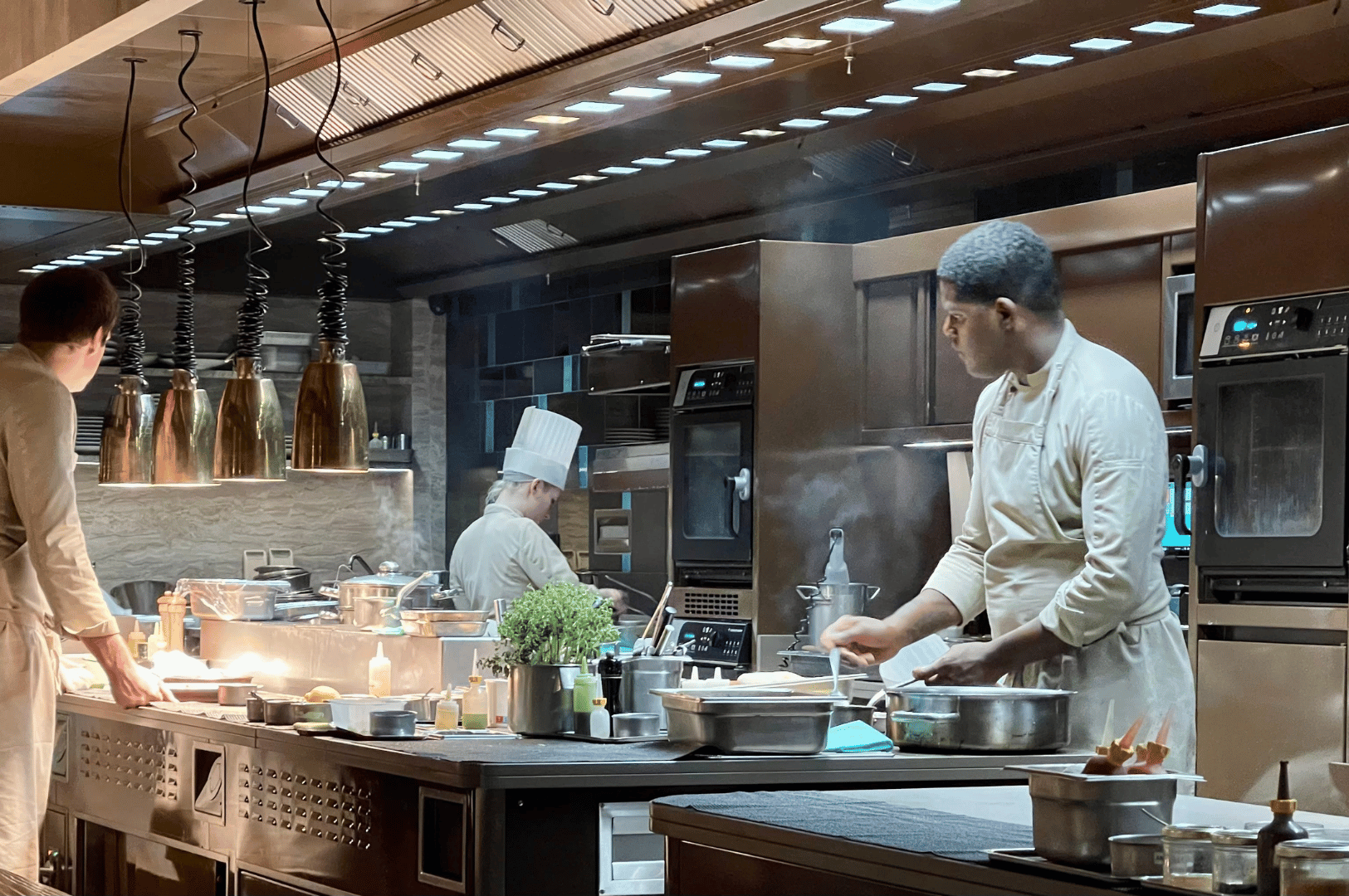 Three chefs work in synchronized motion inside a stainless steel open kitchen in Singapore, surrounded by pots, herbs, and ambient lighting. The dynamic scene captures the precision and intensity of culinary craft, reinforcing the open kitchen as a stage for gastronomic performance.