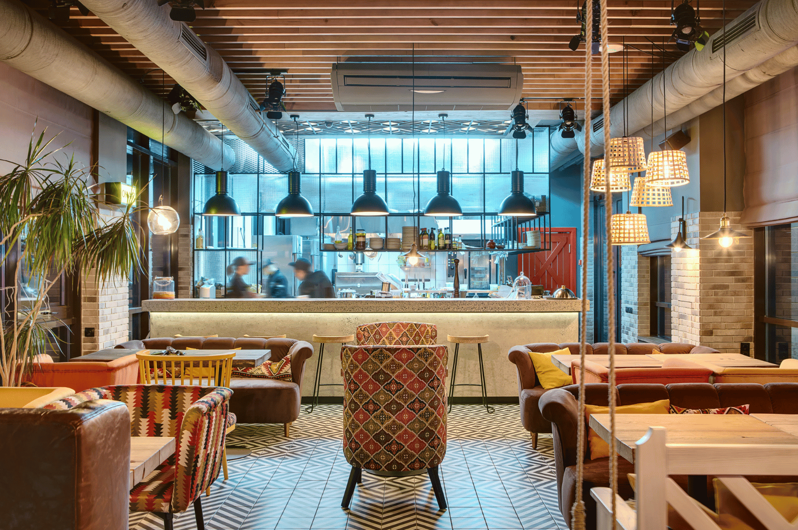 Stylish open-kitchen restaurant in Singapore featuring geometric tile flooring, warm pendant lighting, and eclectic seating with vibrant upholstery. The ambient space blends rustic textures and modern design, while chefs work visibly behind glass, reinforcing the theatrical intimacy of open kitchen architecture.