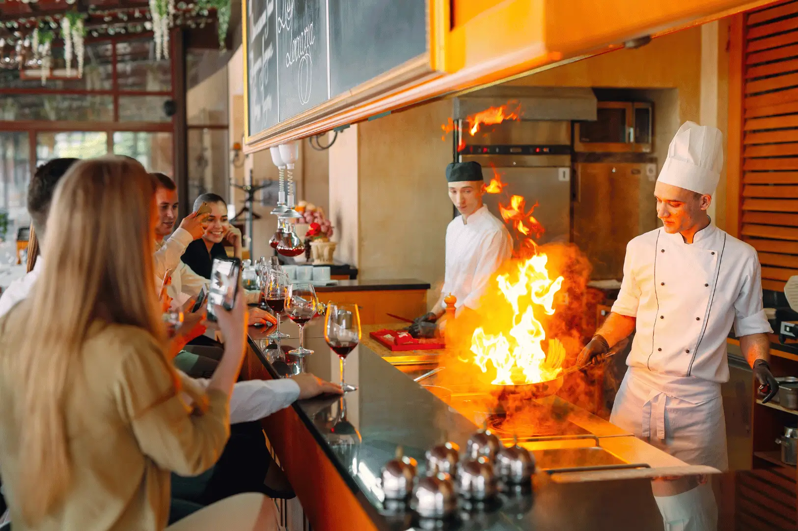 Chef performs a dramatic flambé at a sleek open-kitchen counter in a modern Singapore restaurant, captivating diners seated nearby. Guests sip wine and film the fiery moment, highlighting the immersive, theatrical nature of open kitchen design that blends culinary artistry with guest interaction.