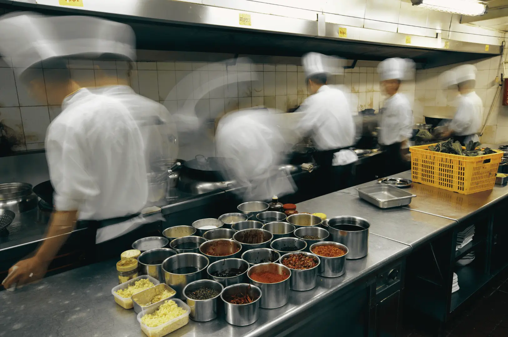 High-energy commercial kitchen in Singapore with chefs in white uniforms working under stainless steel ventilation hoods. Foreground shows a prep counter with spices, chopped garlic, and fresh vegetables, highlighting the heat and humidity challenges addressed by effective kitchen ventilation systems.