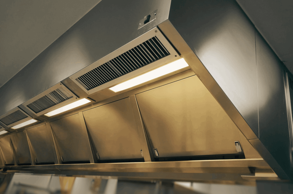 Stainless steel commercial kitchen ventilation hood with angled grease filters and built-in lighting, designed to capture smoke, heat, and airborne grease—illustrating essential airflow solutions for Singapore’s humid culinary environments.