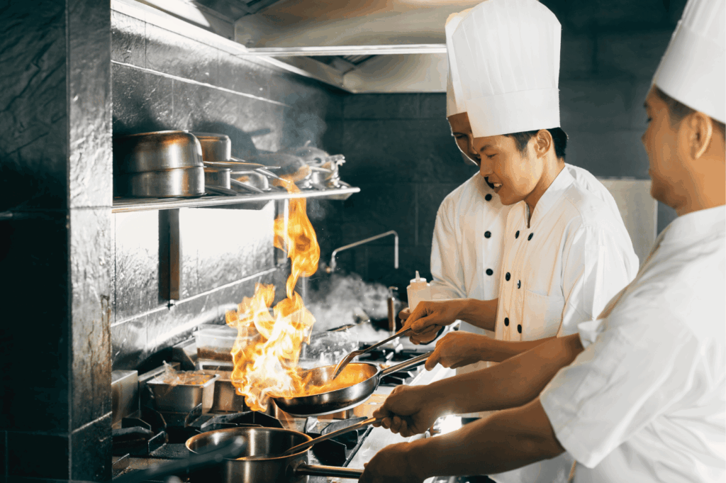 Three chefs in white uniforms and tall hats cooking over open flames in a high-heat commercial kitchen, surrounded by stainless steel equipment and shelving—capturing the dynamic energy and ventilation demands of Singapore’s professional culinary spaces.