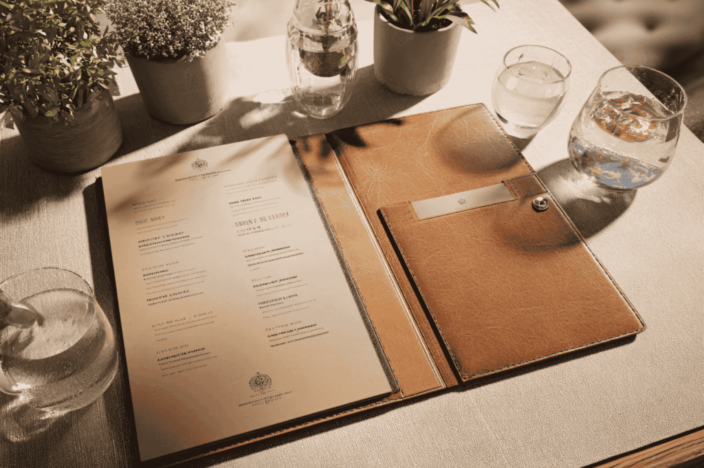 Refined tabletop scene illustrating brand identity through tactile materials and thoughtful presentation; tan leather menu folder, citrus-infused water, and potted plants evoke a sensory-driven hospitality concept rooted in elegance, detail, and curated guest experience.