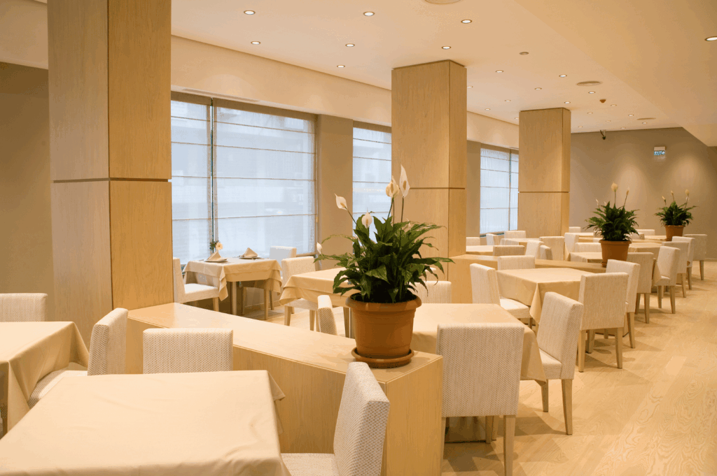 Bright, minimalist dining space embodying brand identity through natural light, soft wood tones, and biophilic accents; peace lilies and beige tablecloths create a serene, welcoming atmosphere that reflects a hospitality concept rooted in calm elegance and organic warmth.