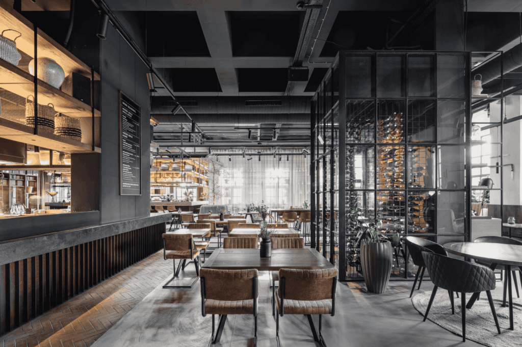 Upscale restaurant interior blending industrial chic with refined elegance; warm wood, metal, and glass elements create a sophisticated ambiance. Leather chairs surround neatly arranged tables, while a tall glass-enclosed wine rack adds vertical drama. The left-side bar features open shelving with baskets and decor, evoking raw materiality. Ambient lighting and natural sunlight from large windows soften the space, highlighting the transformation of raw textures into a polished, inviting dining experience.