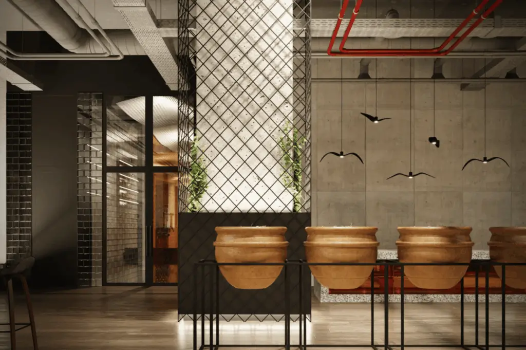Industrial-style restaurant interior blending raw textures with artistic flair; terracotta pots on a black metal stand anchor the foreground, while a mesh-wrapped column glows softly, sprouting greenery at its base. Exposed concrete walls, red piping, and ductwork emphasize the space’s raw architectural bones. Suspended black bird sculptures add a whimsical contrast, and warm wood flooring softens the starkness. A visual narrative of transformation—where industrial grit meets curated design.