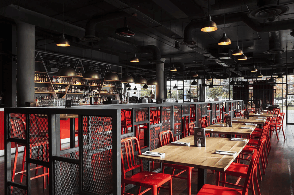 Contemporary industrial restaurant interior featuring rows of red metal chairs paired with wooden tables, each set with menus and napkins. A dark ceiling with exposed ductwork and pendant lights casts warm illumination, while large windows reveal lush greenery outside. The bar area, framed in black metal mesh, anchors the space with stocked shelves and glassware. Raw materials—metal, wood, and glass—are elevated into a stylish, inviting atmosphere, embodying the industrial chic transformation of utilitarian design into refined hospitality.