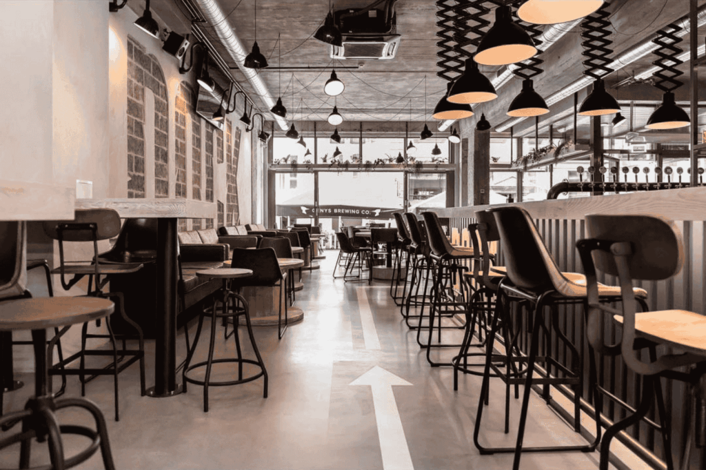 Modern café interior with industrial design elements; black metal chairs and stools surround wooden tables in varied seating arrangements. Exposed ductwork, pendant lights, and a concrete floor reinforce the raw aesthetic, while a white directional arrow adds subtle wayfinding. Brick walls and large windows invite natural light, softened by decorative plants on a nearby shelf. A casual yet curated atmosphere where industrial materials are reimagined for comfort and social connection.