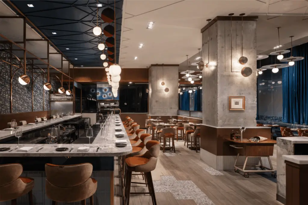 Upscale restaurant interior merging industrial structure with contemporary elegance; exposed concrete pillars and marble countertops contrast with warm wood accents and plush orange-brown seating. Globe pendant lights and recessed ceiling fixtures cast a golden glow, while circular metallic wall art and framed pieces add visual rhythm. Mixed flooring of wood and patterned tile grounds the space in tactile luxury. A refined interpretation of industrial chic—where raw materials are softened into a polished, immersive dining experience.