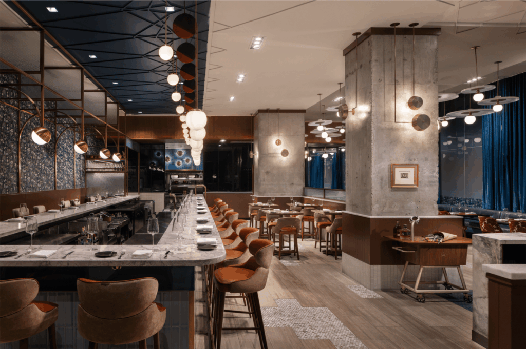Upscale restaurant interior merging industrial structure with contemporary elegance; exposed concrete pillars and marble countertops contrast with warm wood accents and plush orange-brown seating. Globe pendant lights and recessed ceiling fixtures cast a golden glow, while circular metallic wall art and framed pieces add visual rhythm. Mixed flooring of wood and patterned tile grounds the space in tactile luxury. A refined interpretation of industrial chic—where raw materials are softened into a polished, immersive dining experience.