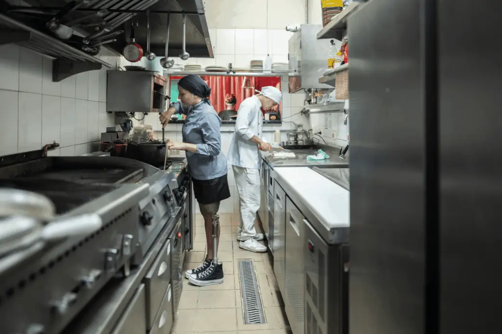 Commercial kitchen in Singapore where a chef with a prosthetic leg prepares food alongside a colleague—highlighting inclusive design, efficient layout, and adaptive workflows that make small kitchens functional and empowering for diverse culinary professionals.
