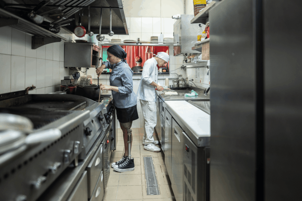 Commercial kitchen in Singapore where a chef with a prosthetic leg prepares food alongside a colleague—highlighting inclusive design, efficient layout, and adaptive workflows that make small kitchens functional and empowering for diverse culinary professionals.