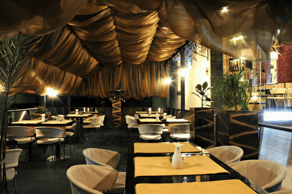 Elegant eco-luxe restaurant interior featuring flowing golden-brown ceiling drapes, white cushioned chairs, and beige placemats. Warm ambient lighting and lush greenery create a refined yet sustainable atmosphere, blending contemporary design with environmentally conscious materials.