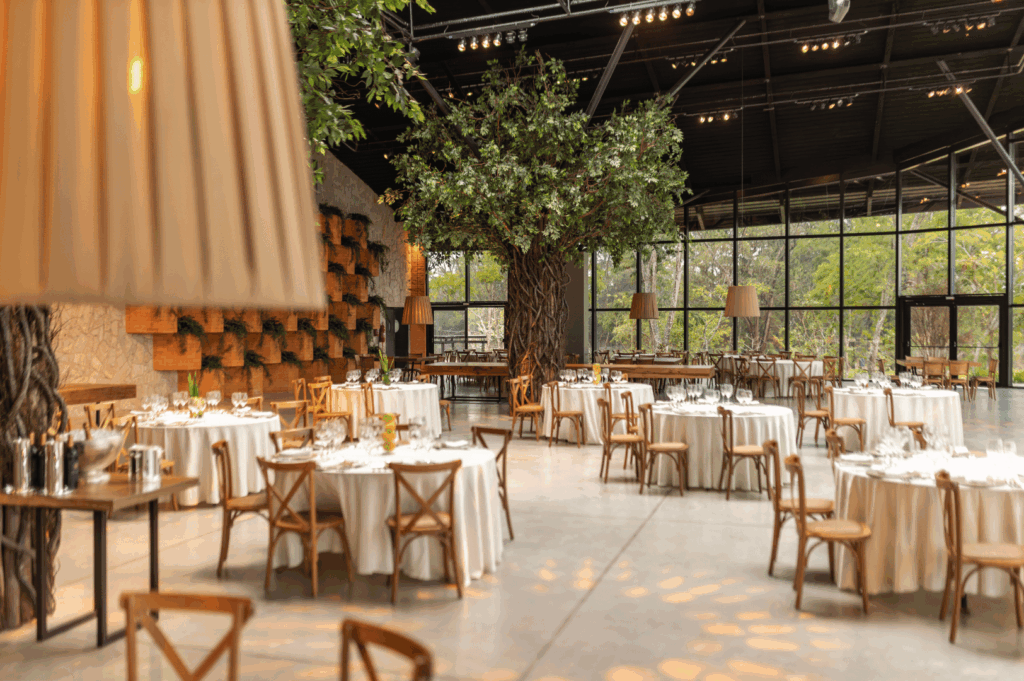 Spacious, light-filled event venue with floor-to-ceiling windows and views of lush greenery. White-clothed round tables and rustic cross-back wooden chairs are arranged beneath warm lighting and whimsical artificial trees, blending natural elegance with sustainable design principles.