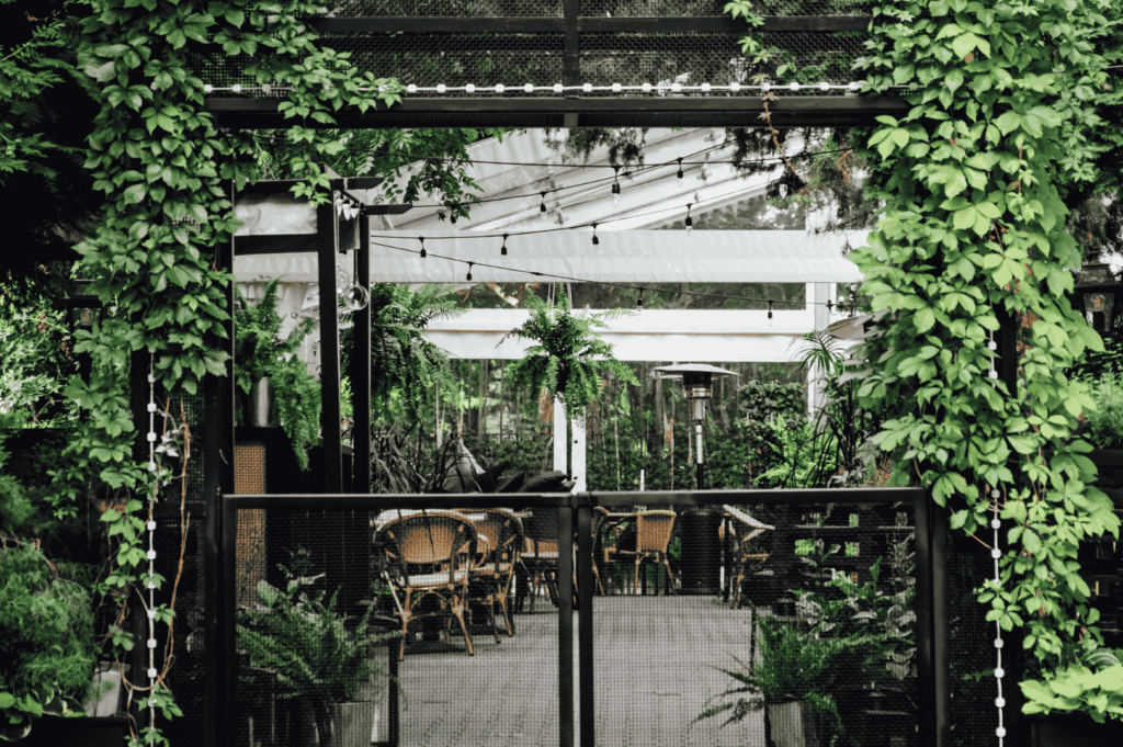 Serene outdoor café entrance framed by a vine-covered black metal archway, leading to a garden-style seating area with wooden tables under a white canopy. Hanging plants and string lights enhance the cozy, nature-integrated ambiance, showcasing sustainable design in harmony with lush greenery.