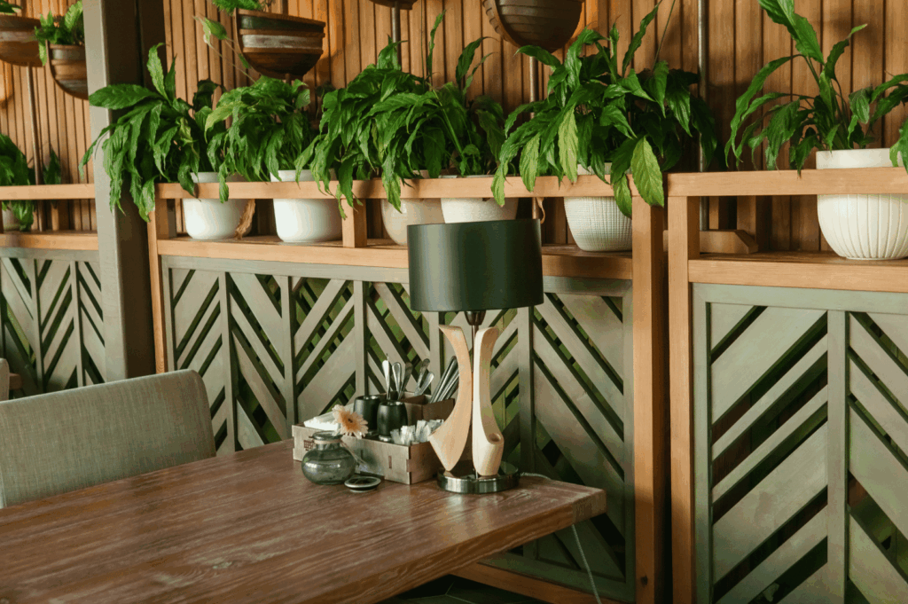 Cozy indoor dining nook featuring a wooden table, cushioned seating, and a geometric wood-paneled wall adorned with potted greenery. A sleek black-and-wood lamp and neatly arranged condiments add modern flair, blending sustainable materials with warm, intimate design.