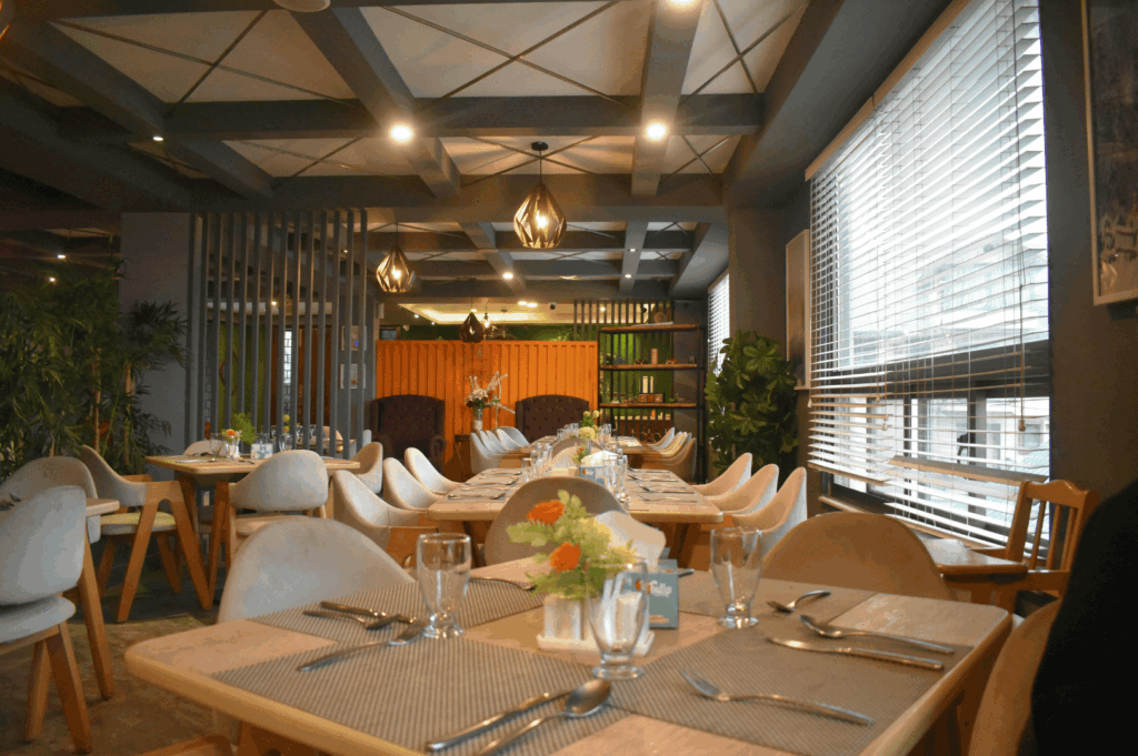 Contemporary restaurant interior with upholstered chairs, wooden tables, and floral centerpieces under geometric ceiling panels and pendant lights. Natural light filters through large windows with horizontal blinds, while wood finishes and greenery add warmth and sustainable elegance to the stylish dining space.