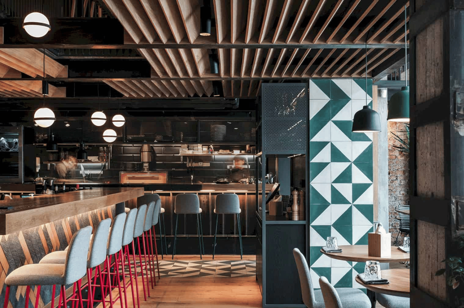 Contemporary Singapore restaurant with open-kitchen layout, featuring a warm wood-accented bar, geometric teal wall tiles, and ambient globe pendant lights. The visible kitchen and cozy seating invite diners into a stylish, interactive space where design and culinary performance converge.
