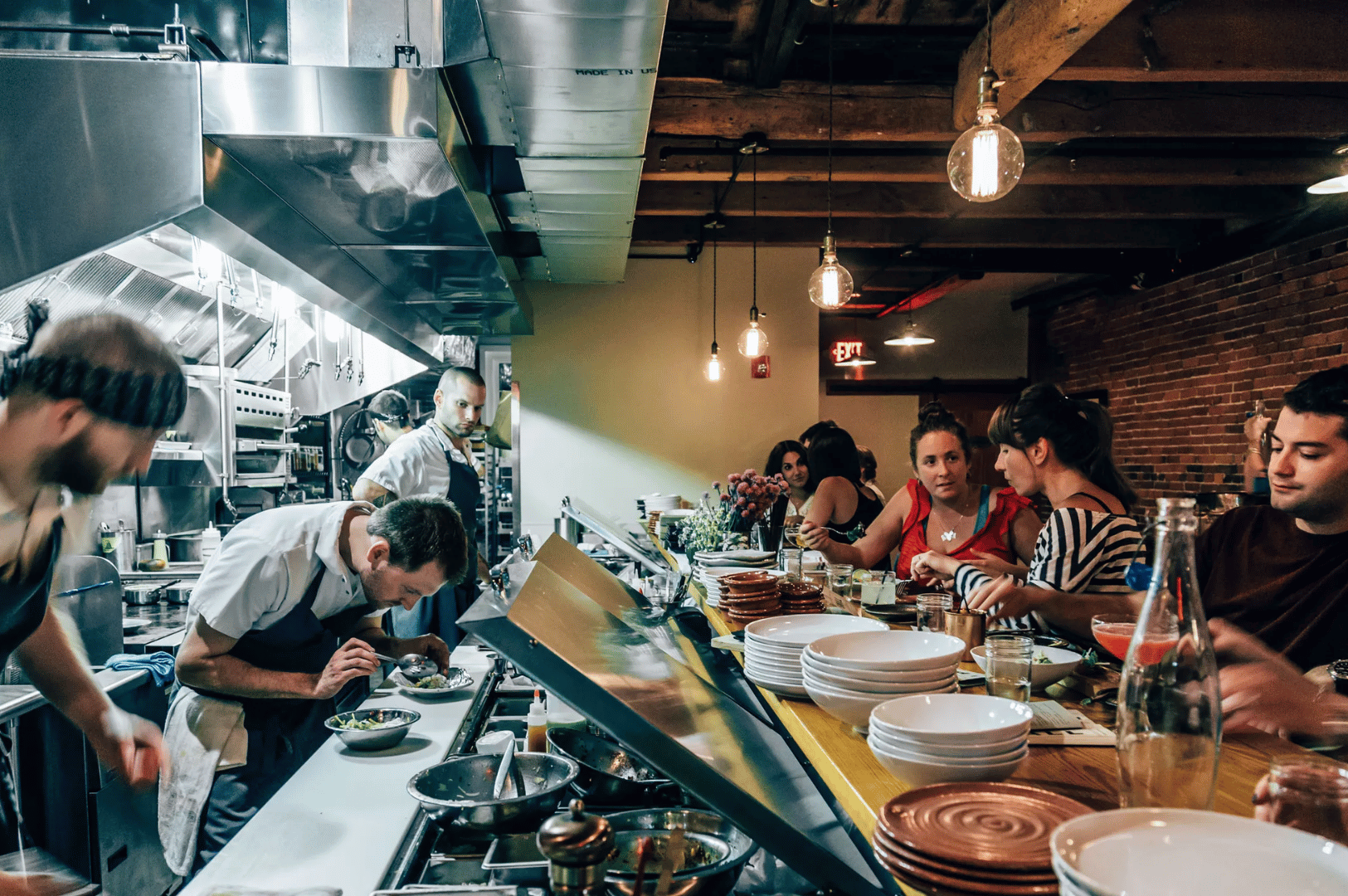 Bustling open kitchen in a Singapore restaurant where chefs plate dishes beside diners seated at a rustic counter. Warm lighting, exposed beams, and stacked serviceware evoke a cozy, communal atmosphere, showcasing how open kitchen design fosters transparency, connection, and shared culinary experience.