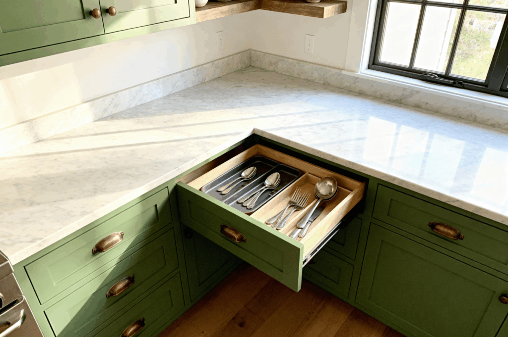 Stylish corner kitchen in Singapore featuring green cabinetry, brass hardware, and a white marble countertop. An open drawer reveals a meticulously organized cutlery tray, exemplifying how thoughtful storage solutions and elegant design can elevate functionality in compact residential spaces.