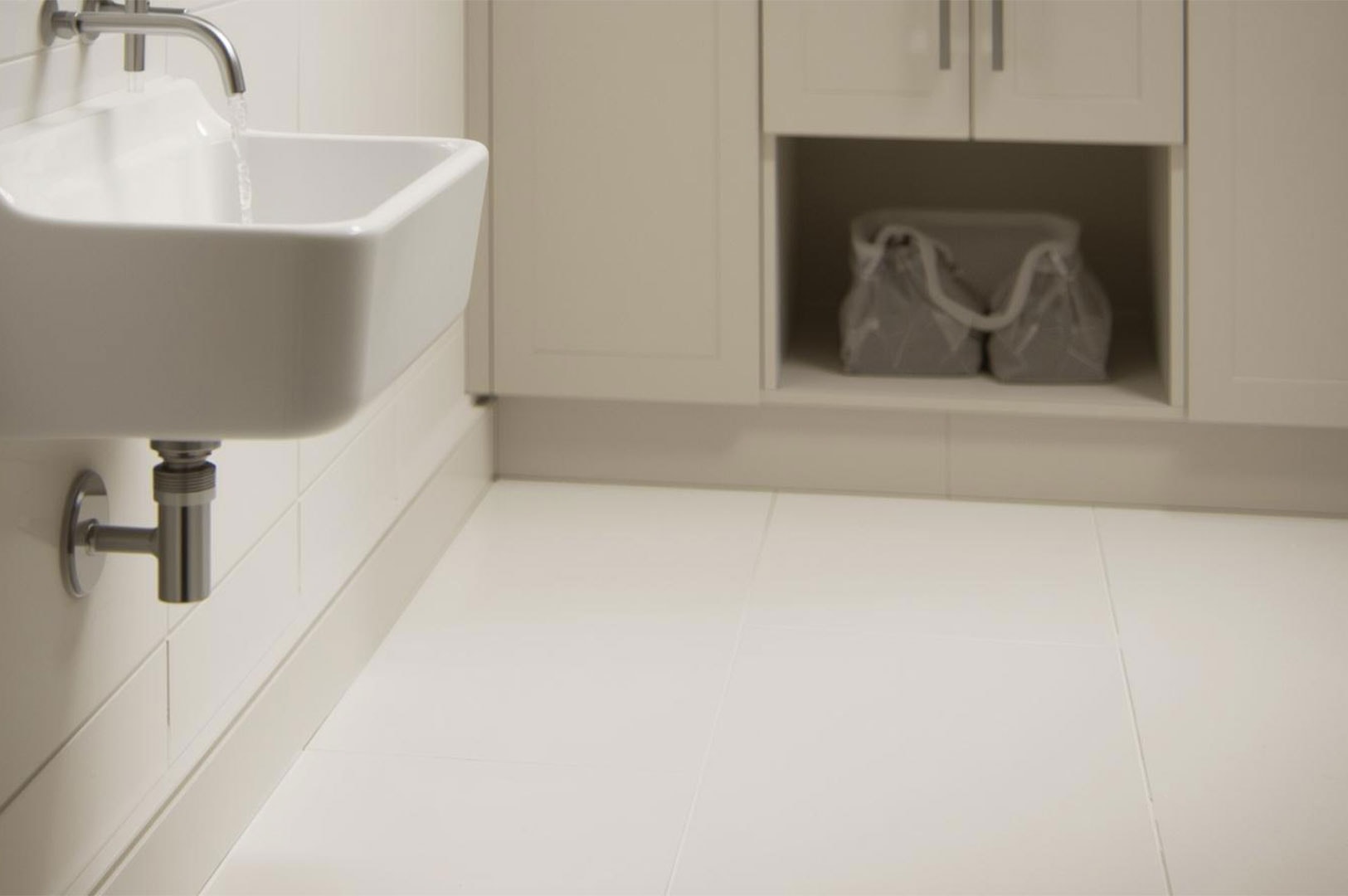 A deep white ceramic utility sink with a wall-mounted chrome faucet and exposed plumbing in a clean, white-tiled laundry or utility room.