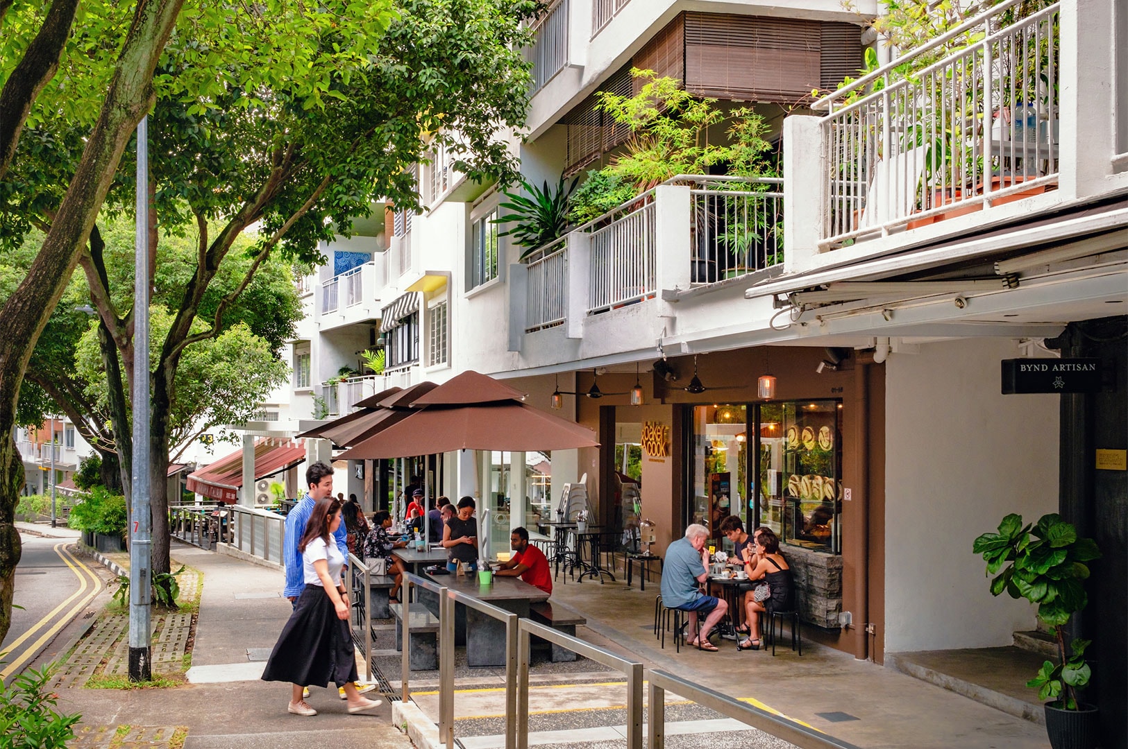 Customers enjoying casual al fresco dining at a sidewalk cafe and bakery named Baker & Cook, located on a sunny, tree-lined street with apartment buildings above.