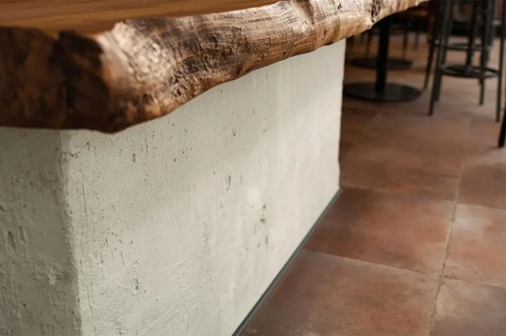 Close-up architectural detail of a bar counter featuring a live-edge wood top sitting on a rough textured white concrete base.