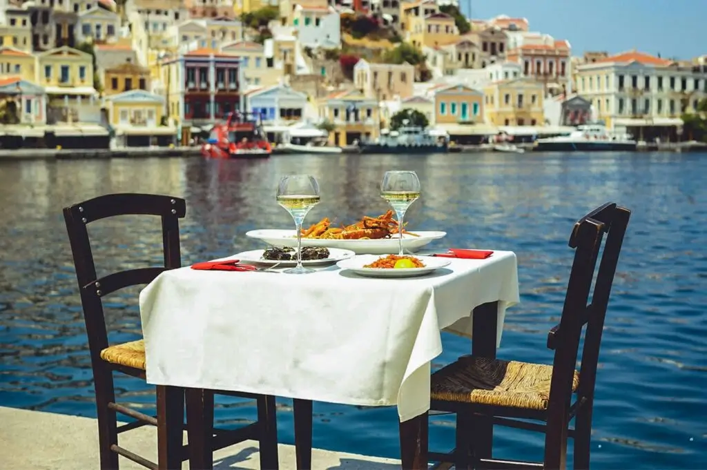 Romantic waterfront dining table set with white wine and seafood, overlooking a sunny, colorful Greek harbor village.