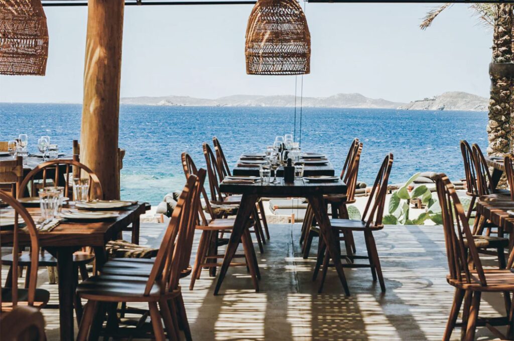 Open-air bohemian restaurant setting with rustic wooden furniture and large woven wicker pendant lights framing a view of the blue ocean.