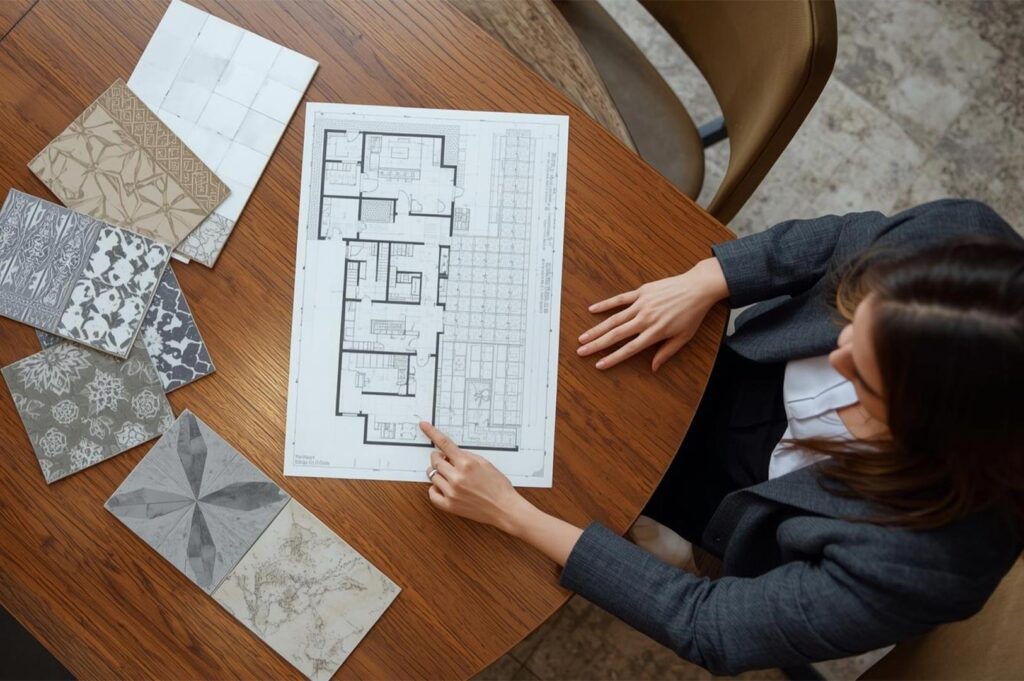 Overhead view of an interior designer pointing at an architectural floor plan while reviewing various patterned bathroom tile samples on a wooden table.