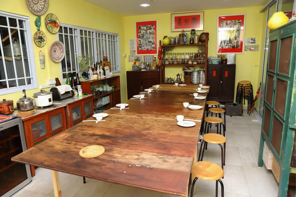 A long rustic wooden dining table set with white bowls and stools in a vibrant yellow room featuring traditional Chinese decor and shelving.