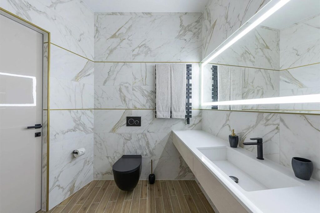 A modern luxury bathroom interior featuring white marble walls with gold veining, a floating vanity, a backlit LED mirror, and a wall-mounted black toilet.