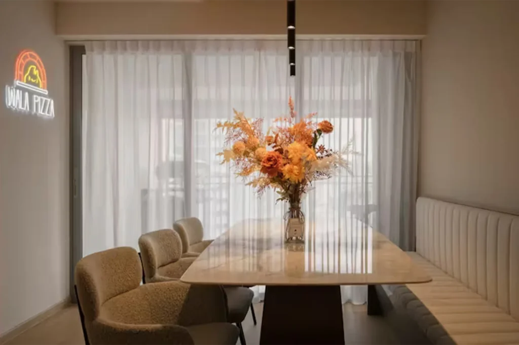 A chic modern dining area with a marble table, plush beige chairs, a large autumn floral arrangement, and a "Wala Pizza" neon sign on the wall.
