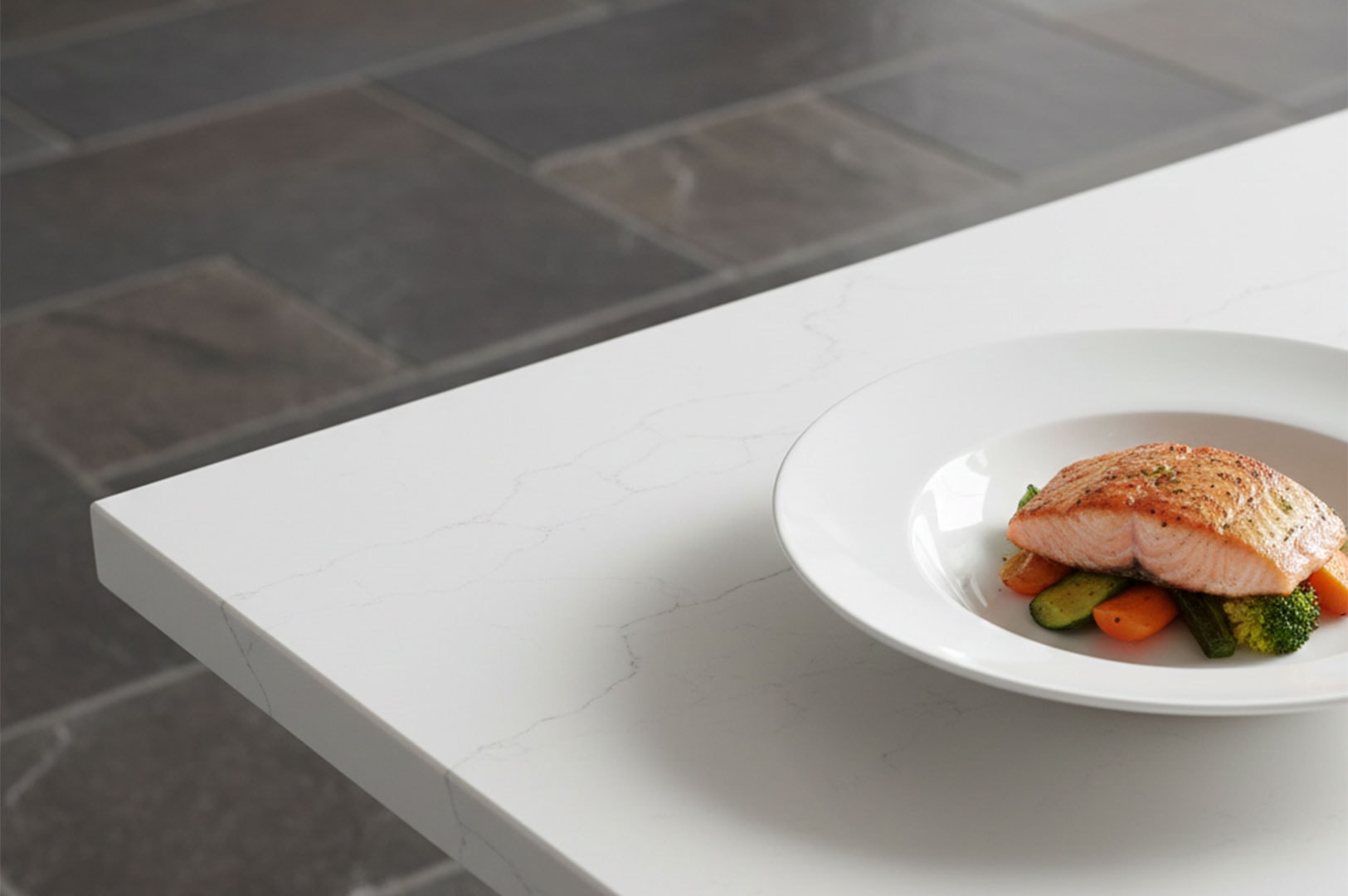 A gourmet dish featuring a grilled salmon fillet served over roasted carrots and broccoli on a white plate, placed on a sleek white stone table with a slate floor background.