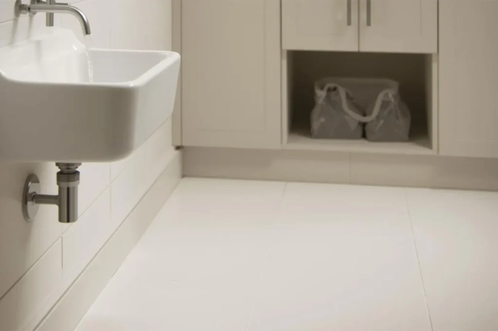 A deep white ceramic utility sink with a wall-mounted chrome faucet and exposed plumbing in a clean, white-tiled laundry or utility room.
