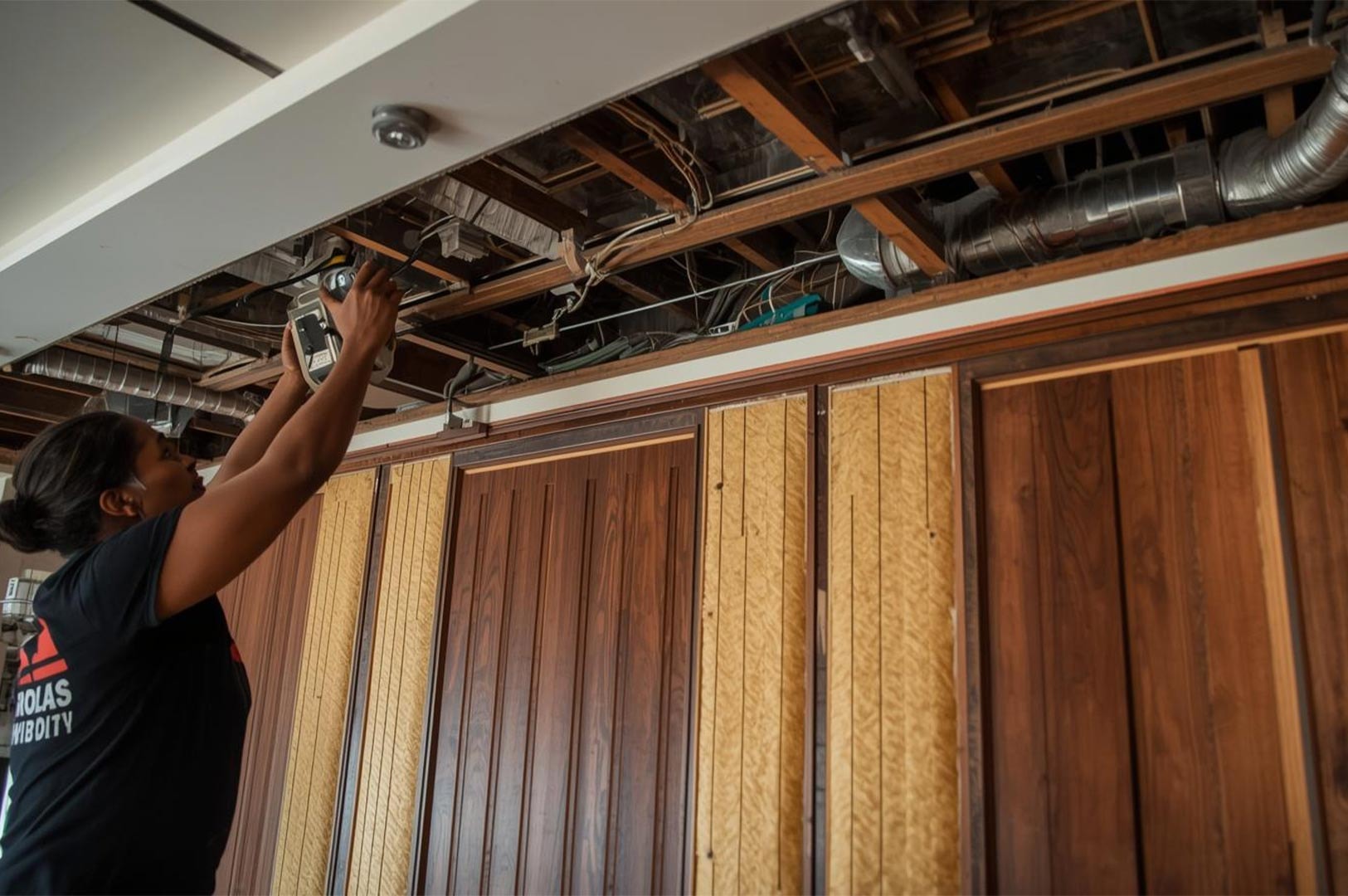 A construction worker installing HVAC ductwork and electrical wiring in an exposed ceiling above wooden wall paneling during a commercial renovation project.