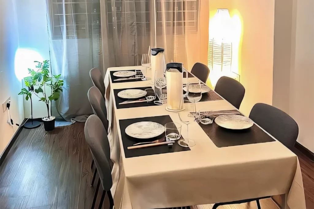 A sophisticated long dining table set with black placemats, white plates, and wine glasses, illuminated by soft warm ambient floor lamps.