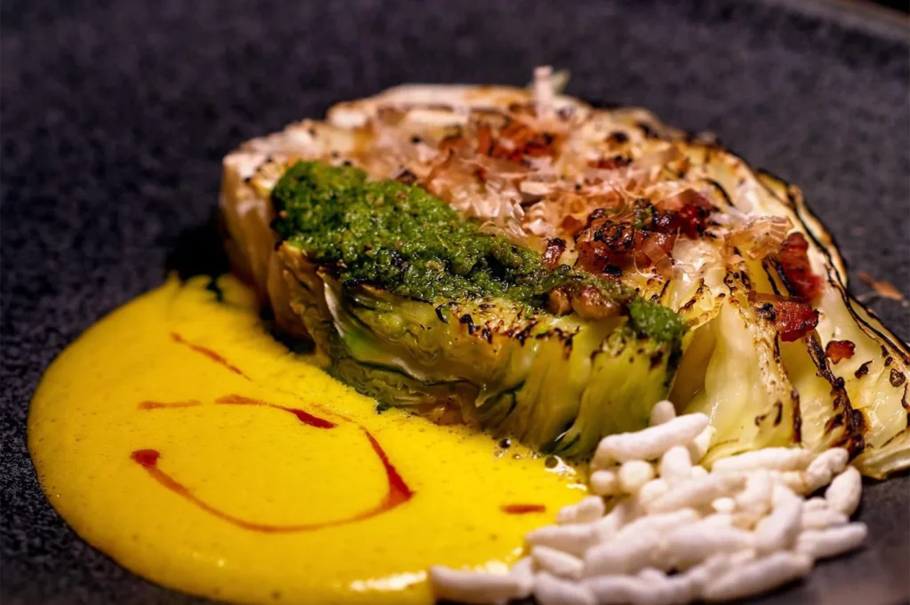 Close-up of a gourmet plated dish featuring charred cabbage topped with green pesto and bonito flakes, served with a yellow carrot puree and puffed rice.