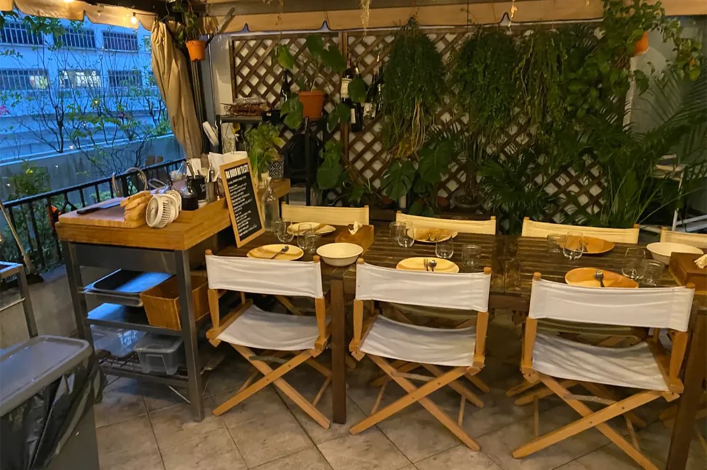 An alfresco dining setup on a balcony featuring wooden tables, white canvas director chairs, and a lush green plant wall with lattice fencing.
