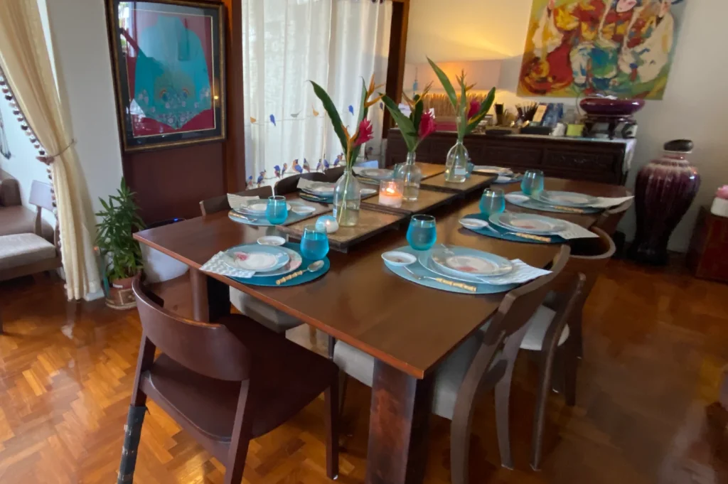 A warm wooden dining table set for a group, accented by bright blue glassware, tropical floral centerpieces, and bold contemporary artwork on the walls.