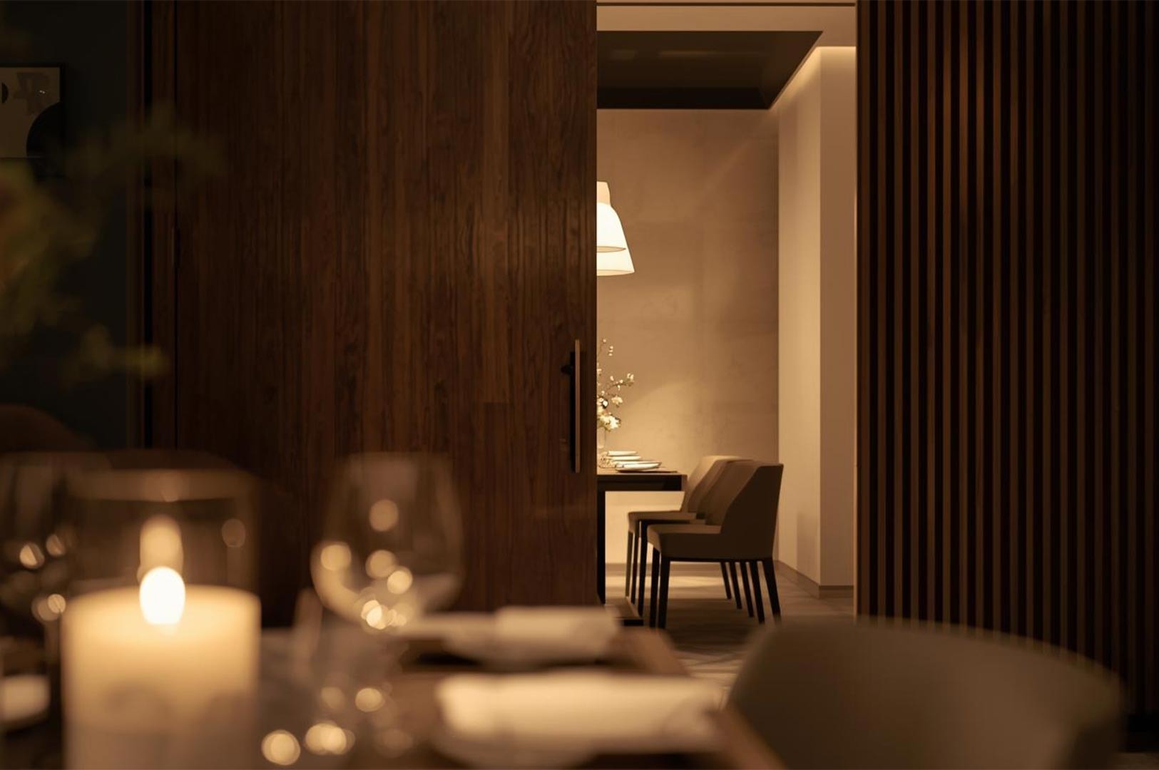 View through a partially open dark wooden sliding door revealing a modern, minimalist dining room with warm ambient lighting and a set table.