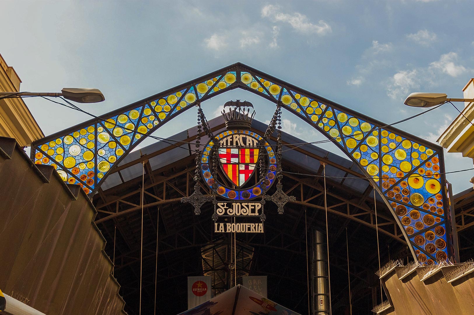 The ornate Art Nouveau iron entrance of the Mercat de Sant Josep de la Boqueria in Barcelona, featuring colorful stained glass circles and the iconic city crest under a blue sky.