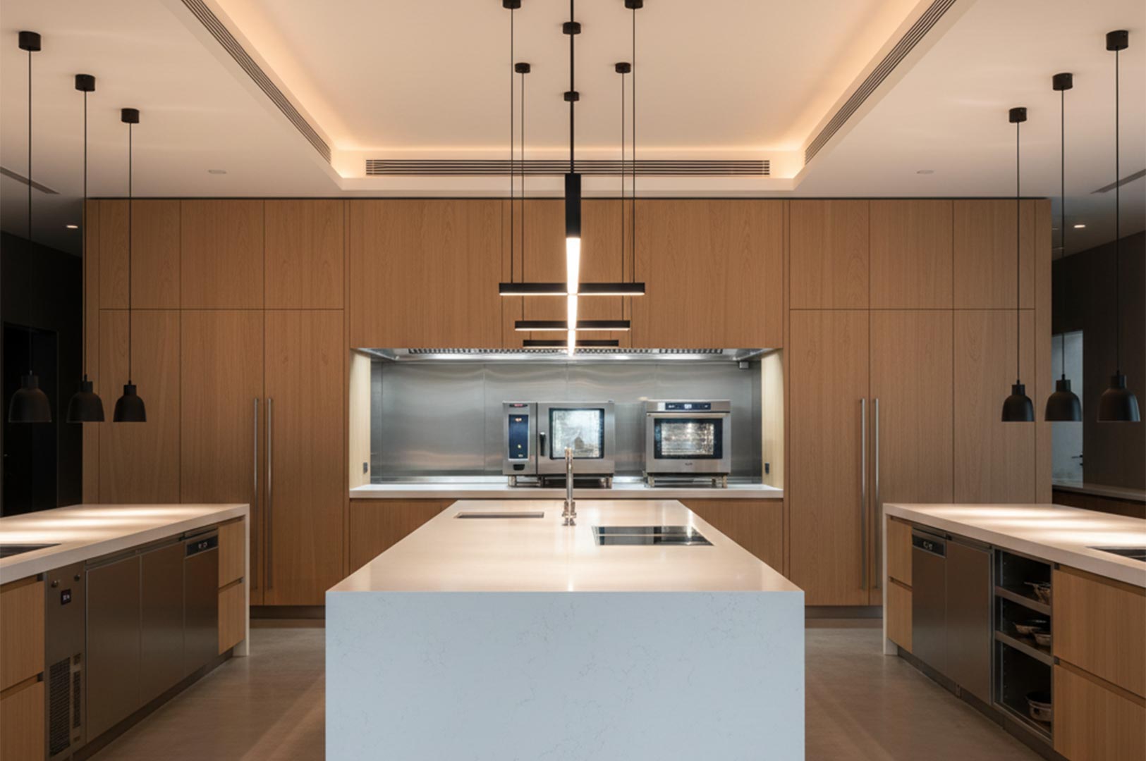 A symmetrical view of a high-end commercial kitchen featuring floor-to-ceiling light wood cabinetry, a large white marble island with a built-in cooktop, and modern black linear pendant lighting.