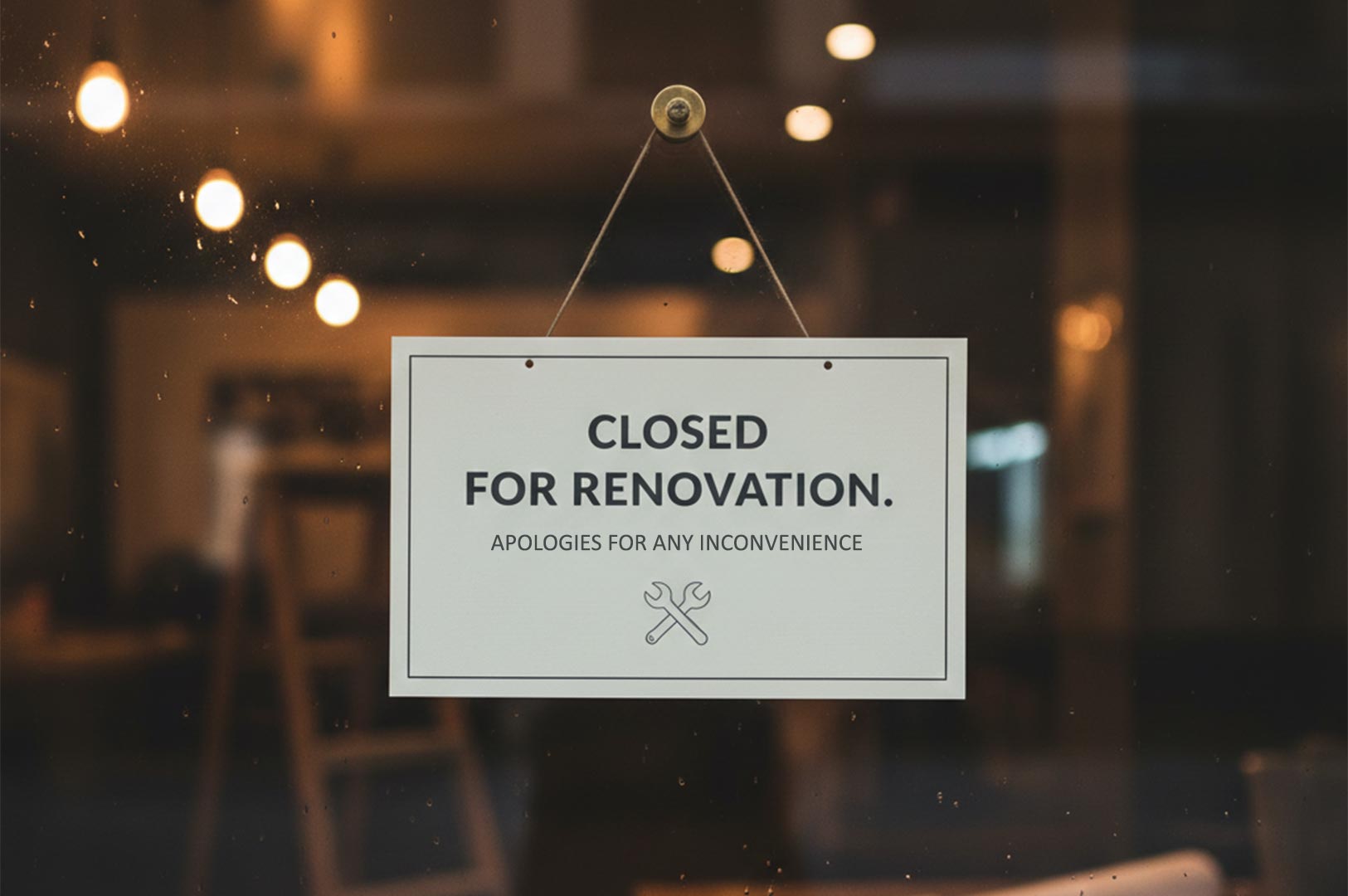 A white rectangular sign hanging on a glass door that reads "CLOSED FOR RENOVATION" with a wrench icon, showing a blurred interior background.